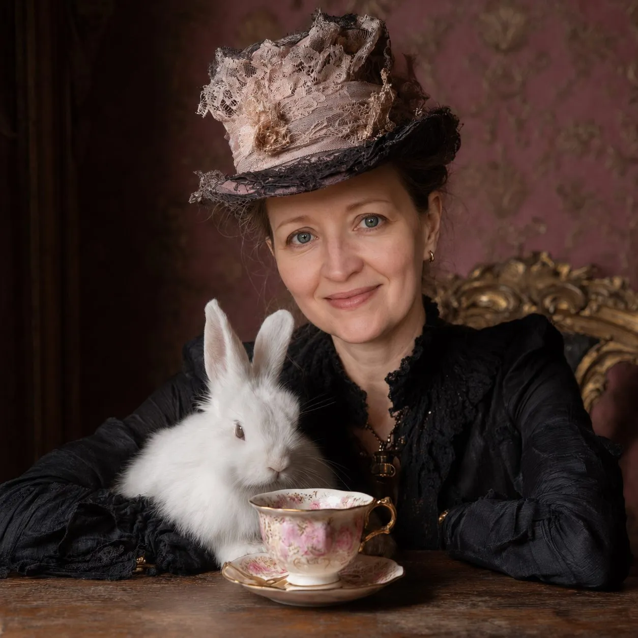 Diane Whiddon in a lace-trimmed Victorian hat, seated beside a white rabbit and a pink floral teacup against rose damask wallpaper